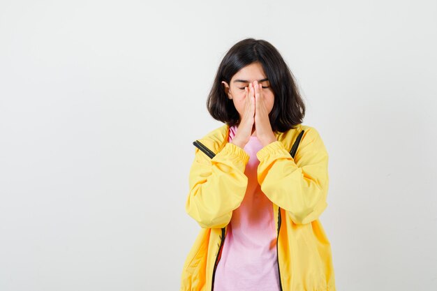 Chica adolescente en camiseta, chaqueta amarilla cogidos de la mano en gesto de oración y mirando enfocado, vista frontal.