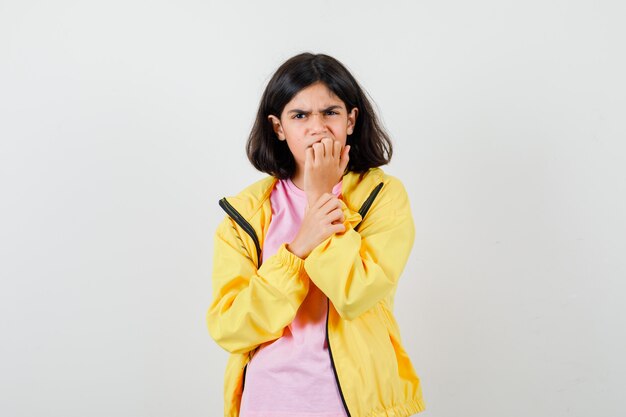 Chica adolescente en camiseta, chaqueta amarilla mordiendo las uñas y mirando estresado, vista frontal.