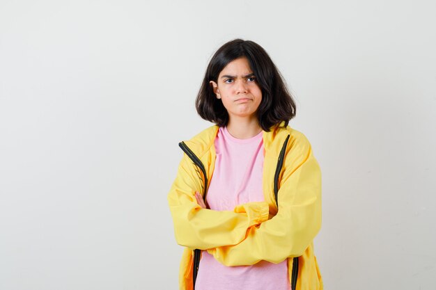 Chica adolescente en camiseta, chaqueta amarilla sosteniendo los brazos cruzados y mirando molesto, vista frontal.