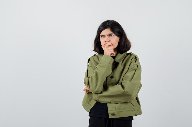 Chica adolescente en camiseta, chaqueta apoyando la barbilla en la mano y mirando pensativo, vista frontal.