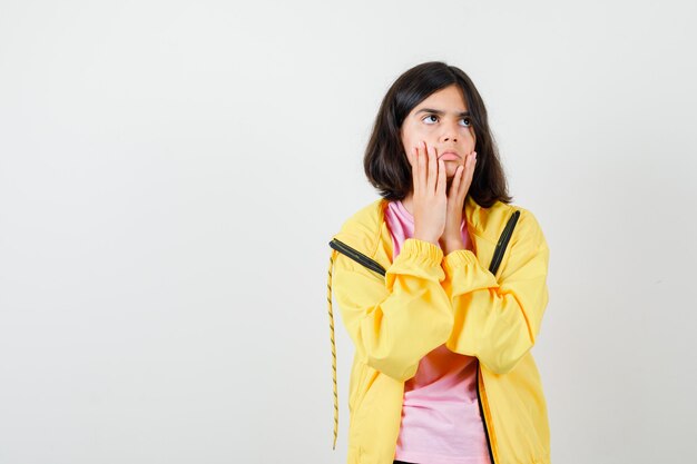 Chica adolescente en camiseta, chaqueta cogidos de la mano cerca de la boca y mirando atenta, vista frontal.