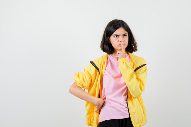 Chica adolescente en camiseta, chaqueta mostrando gesto de silencio, mirando a otro lado y mirando enfocado, vista frontal.