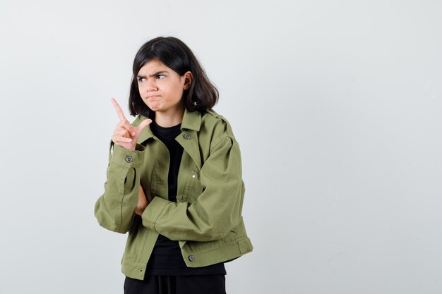 Chica adolescente en camiseta, chaqueta verde apuntando hacia afuera y mirando serio, vista frontal.