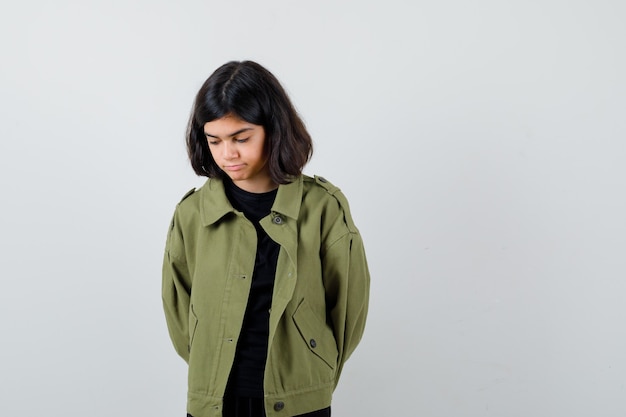 Chica adolescente en camiseta, chaqueta verde cogidos de la mano detrás de la espalda mientras mira hacia abajo y mira pensativa, vista frontal.