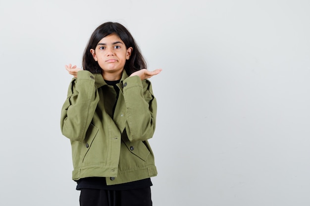 Chica adolescente en camiseta, chaqueta verde mostrando gesto de duda y mirando indeciso, vista frontal.
