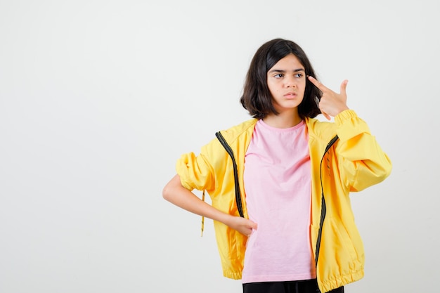 Chica adolescente en chándal amarillo, camiseta apuntando a las sienes con el dedo y mirando insatisfecho, vista frontal.