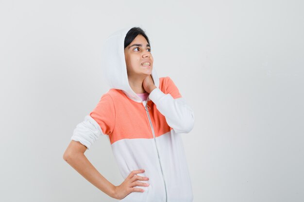 Chica adolescente con chaqueta blanca sosteniendo la mano en su cuello mientras mira a un lado y parece nervioso.