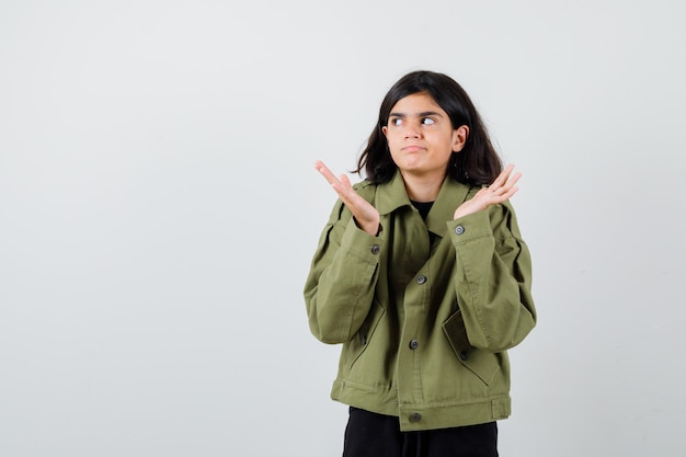 Chica adolescente en chaqueta verde militar mostrando gesto de impotencia, mirando hacia arriba y mirando confundido, vista frontal.