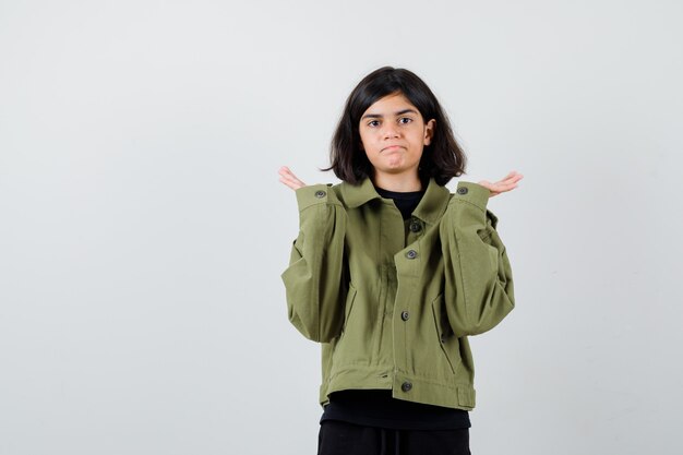 Chica adolescente en chaqueta verde militar mostrando gesto de impotencia y mirando perplejo, vista frontal.