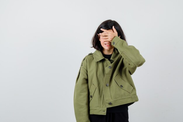 Chica adolescente en chaqueta verde militar sosteniendo la mano en los ojos y mirando emocionado, vista frontal.