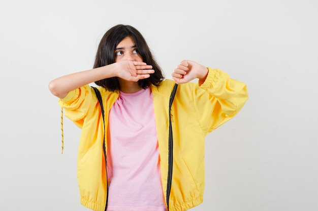 Chica adolescente cubriendo la boca con la mano en chándal amarillo, camiseta y mirando perplejo, vista frontal.