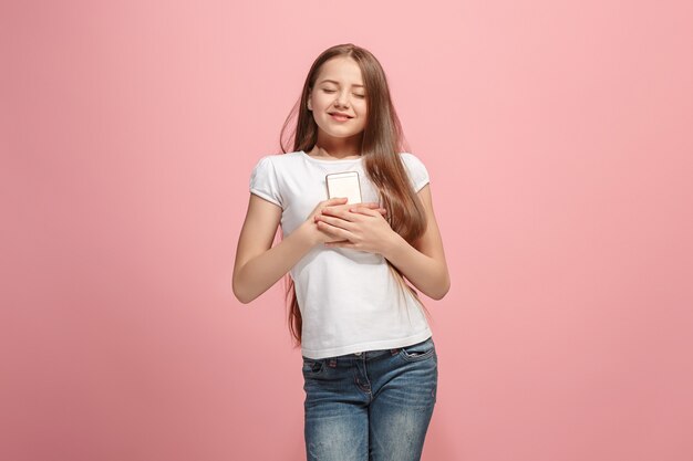 La chica adolescente feliz con teléfono de pie y sonriendo contra la pared rosa