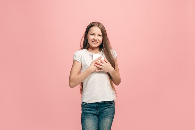 La chica adolescente feliz con teléfono de pie y sonriendo contra la pared rosa