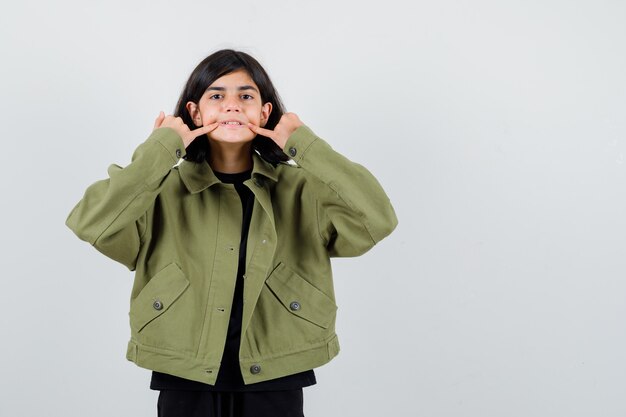 Chica adolescente fuerza una sonrisa en la cara en camiseta, chaqueta verde y mirando enfocado, vista frontal.