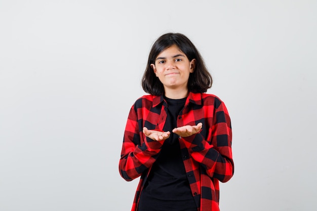 Chica adolescente haciendo gesto de dar o recibir en camiseta, camisa a cuadros y mirando complacido, vista frontal.