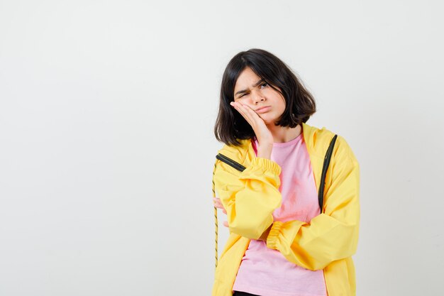 Chica adolescente manteniendo la mano en la mejilla en camiseta, chaqueta y mirando serio, vista frontal.