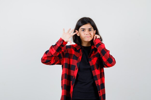 Chica adolescente mirando a la cámara mientras muestra la palma en la camiseta, camisa a cuadros y mirando confiado, vista frontal.