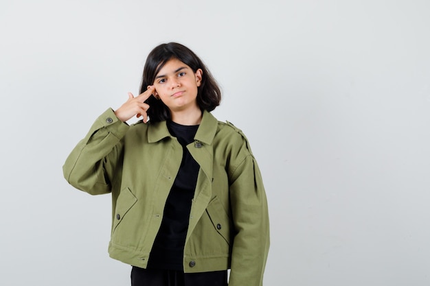 Chica adolescente mostrando gesto de pistola en camiseta, chaqueta verde y mirando seguro de sí mismo, vista frontal.