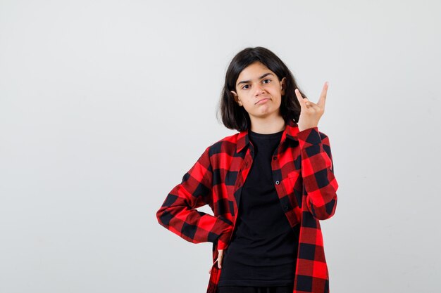 Chica adolescente mostrando gesto de rock en camiseta, camisa a cuadros y mirando seguro de sí mismo, vista frontal.