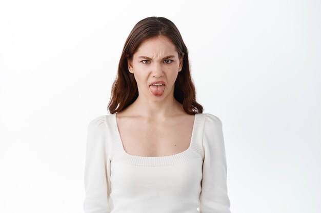 Chica adolescente mostrando la lengua de disgusto y aversión, algo de mal sabor, frunciendo el ceño y quejándose, mirando disgustado, de pie contra la pared blanca