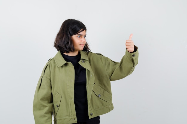 Chica adolescente mostrando el pulgar hacia arriba en camiseta, chaqueta verde y mirando complacido. vista frontal.