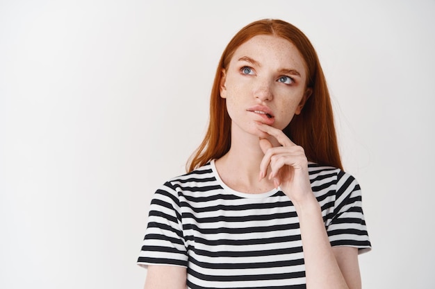 Chica adolescente pensativa con pecas y cabello rojo mordiendo el labio, frunciendo el ceño y mirando hacia arriba mientras piensa, toma una decisión, de pie sobre una pared blanca