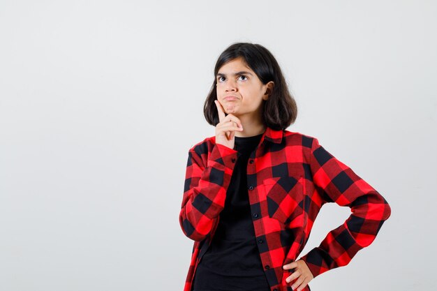 Chica adolescente de pie en pose de pensamiento en camiseta, camisa a cuadros y mirando pensativo. vista frontal.