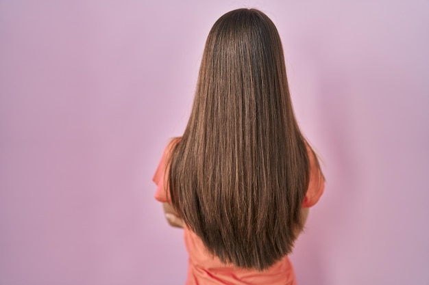 Chica adolescente de pie sobre fondo rosa de pie hacia atrás mirando hacia otro lado con los brazos cruzados