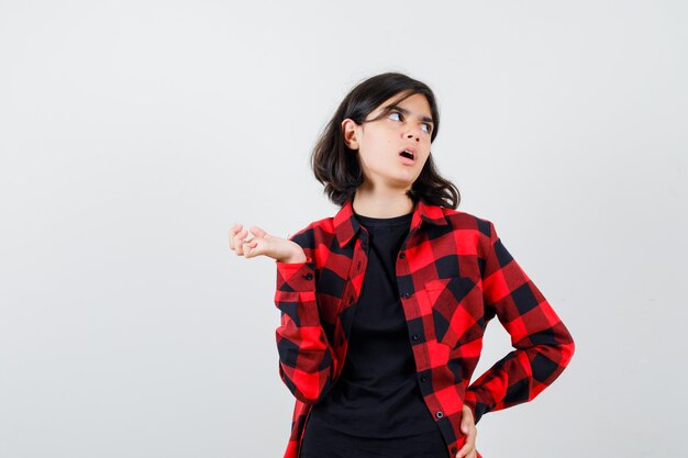 Chica adolescente posando mientras mira a un lado, extendiendo la palma hacia un lado en camiseta, camisa a cuadros y mirando perplejo, vista frontal.