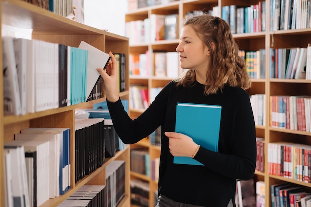 Chica adolescente recogiendo un libro en un estante de la biblioteca