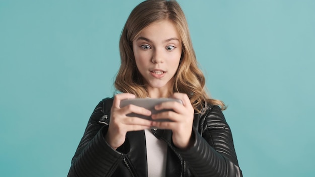 Chica adolescente rubia emocionada jugando en el teléfono inteligente obsesionada con los juegos en línea que se ve nerviosa por el fondo azul