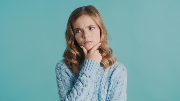 Chica adolescente rubia pensativa mirando preocupación manteniendo la mano en la barbilla pensando en algo sobre fondo azul Expresión facial