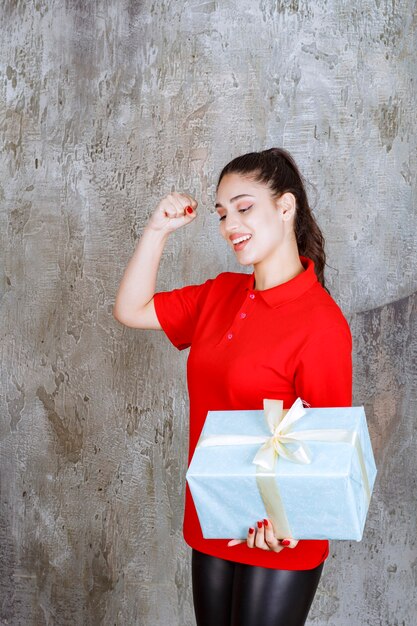 Chica adolescente sosteniendo una caja de regalo azul envuelta con cinta blanca y mostrando el signo de la mano de disfrute.