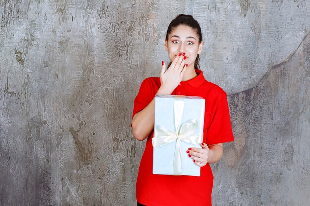 Chica adolescente sosteniendo una caja de regalo azul envuelta con cinta blanca y parece estresada o aterrorizada