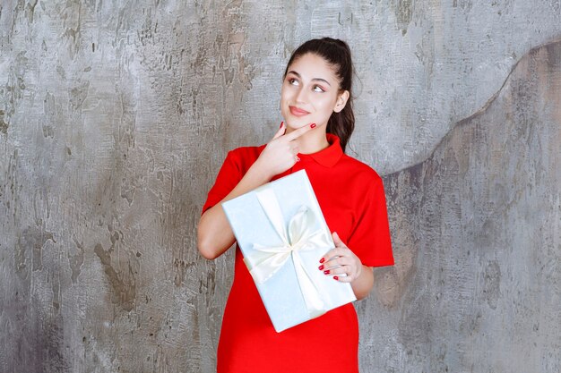 Chica adolescente sosteniendo una caja de regalo azul envuelta con cinta blanca y soñando o teniendo una buena idea