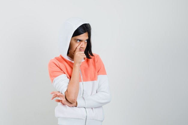 Chica adolescente sosteniendo el dedo en la mejilla en una sudadera y mirando perdido.