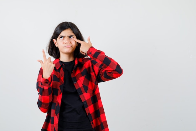 Chica adolescente sosteniendo el dedo en el párpado mientras apunta hacia arriba en una camiseta, camisa a cuadros y mirando pensativo, vista frontal.