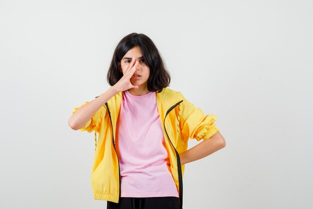 Chica adolescente sosteniendo la mano en la cara, manteniendo la mano en la cintura en camiseta, chaqueta y mirando pensativo, vista frontal.