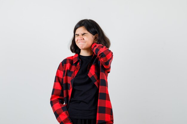 Chica adolescente sosteniendo la mano detrás del cuello en camiseta, camisa a cuadros y mirando doloroso, vista frontal.