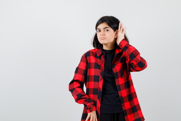 Chica adolescente sosteniendo la mano detrás de la oreja en camiseta, camisa a cuadros y mirando con cuidado, vista frontal.