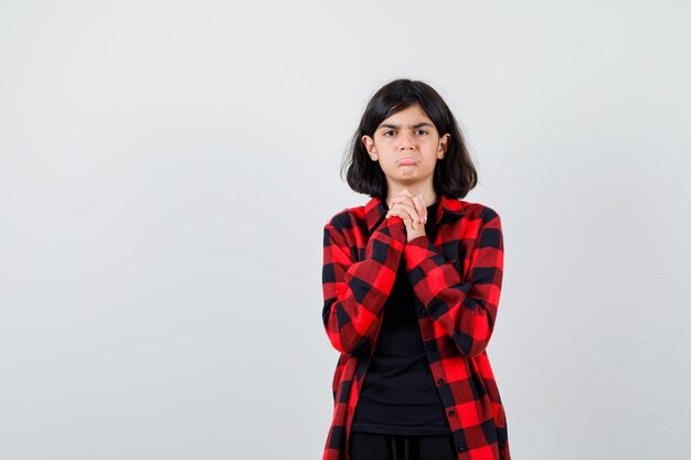 Chica adolescente sosteniendo las manos juntas en gesto de súplica, curvando el labio inferior en camisa casual y mirando gruñón, vista frontal.