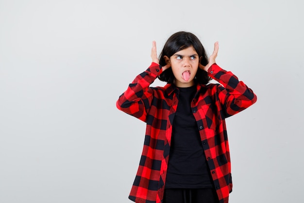 Foto gratuita chica adolescente tapando los oídos con los dedos, sacando la lengua en una camisa casual y luciendo juguetona, vista frontal.