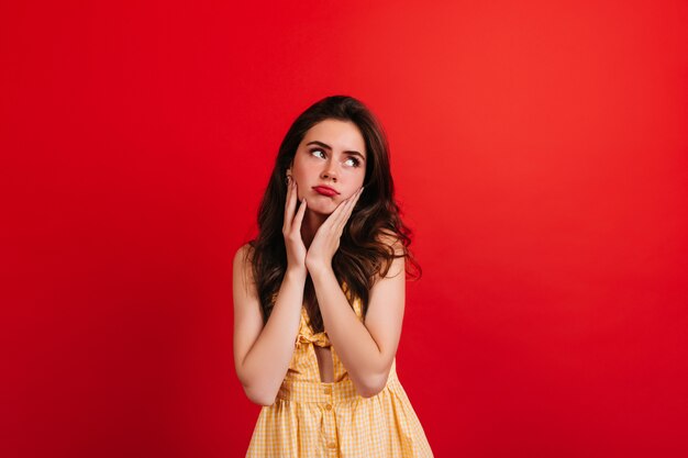 Chica adolescente en vestido amarillo posando de humor triste contra la pared roja. Closeup retrato de mujer morena con labios rojos.
