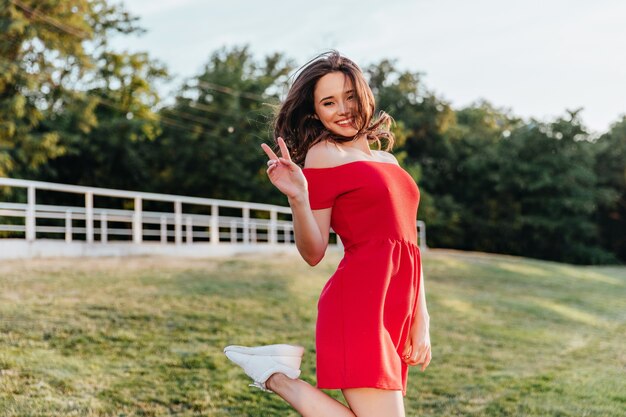 Chica adorable bien formada en traje rojo posando en el parque con placer. Espectacular mujer morena vestida de pie en la naturaleza con el signo de la paz.