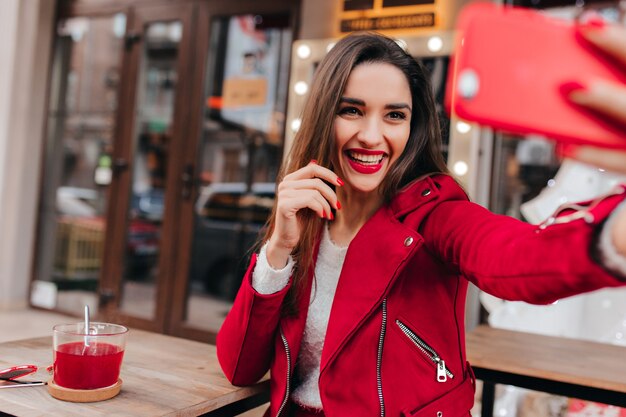 Chica adorable con bonita sonrisa sosteniendo teléfono rojo y haciendo selfie