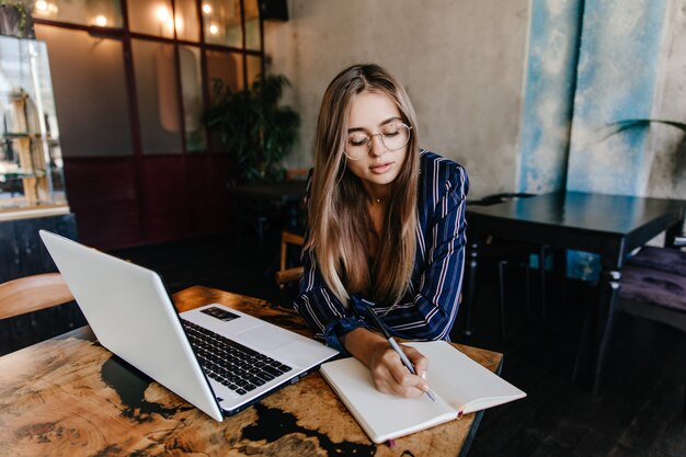 Chica adorable escribiendo algo en su cuaderno. Filmación en interiores de adorable mujer de pelo largo con portátil blanco en café.