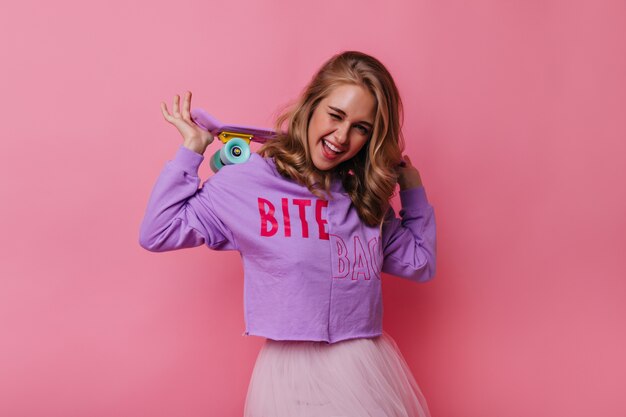 Chica adorable con linda patineta rosa sonriendo a la cámara. Retrato de modelo femenino interesado en camisa morada.