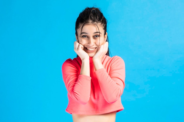 Una chica adorable se llevó las manos a la barbilla y sonrió a la cámara con fondo azul Foto de alta calidad