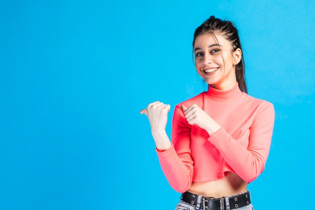 Una chica adorable parada en un fondo azul y señalando sus pulgares a un lado