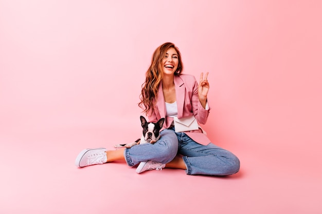 Chica adorable posando con el signo de la paz sentada en el suelo con su perro. Retrato de mujer complacida pelirroja jugando con cachorro.